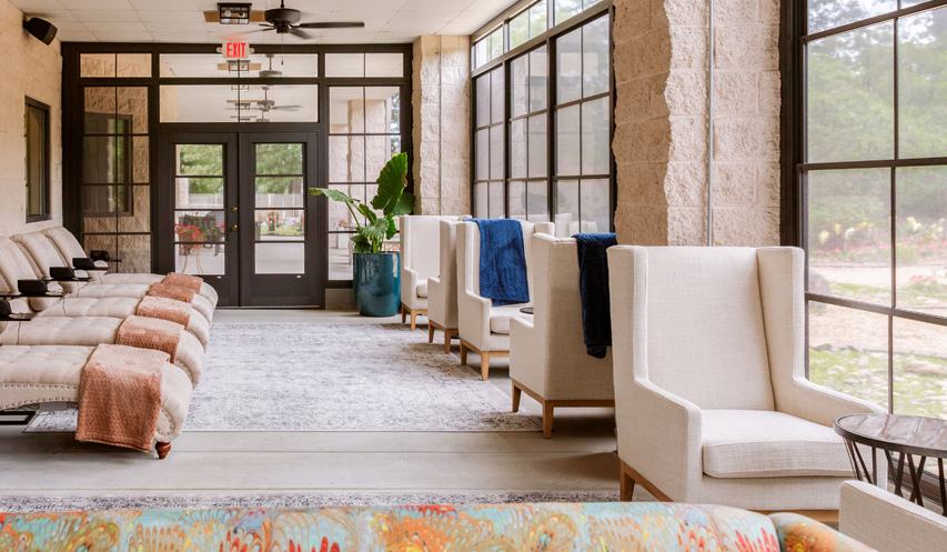 - The Renaissance Day Spa & Alternative Wellness, Fayetteville NC A row of beige lounge chairs with blankets faces large windows in a sunlit room with potted plants, rugs, and high-back armchairs near the entrance. - The Renaissance Day Spa & Alternative Wellness, Fayetteville NC